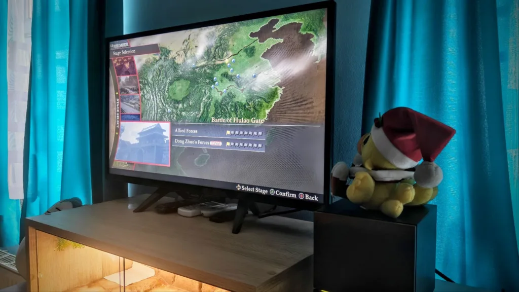 Zrzut ekranu przedstawiający wybór poziomu Battle of Hulao Gate w grze Dynasty Warriors na telewizorze stojącym na szafce.