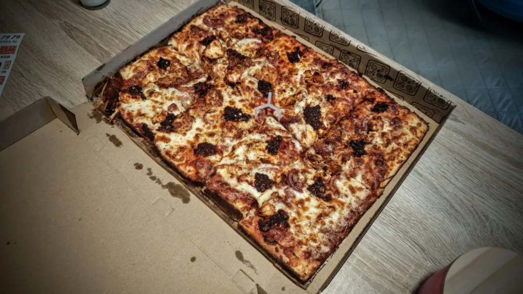 Prostokątną pizzę podzieloną na kawałki w kartonowym pudełku z elementem dystansującym po środku.