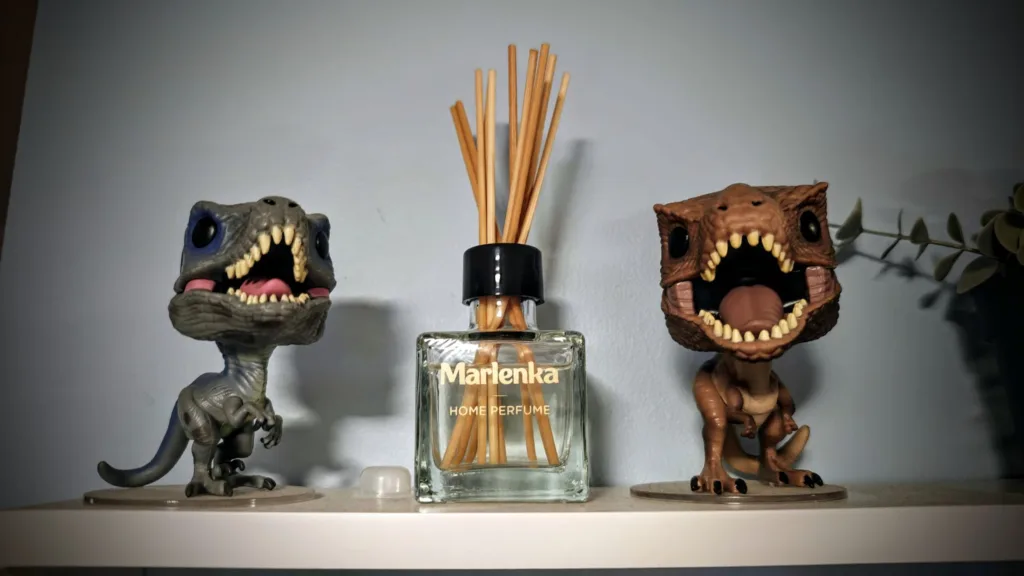 Dwie figurki dinozaurów Funko Pop stoją po bokach flakonu odświeżacza powietrza Marlenka Home Perfume z patyczkami zapachowymi.