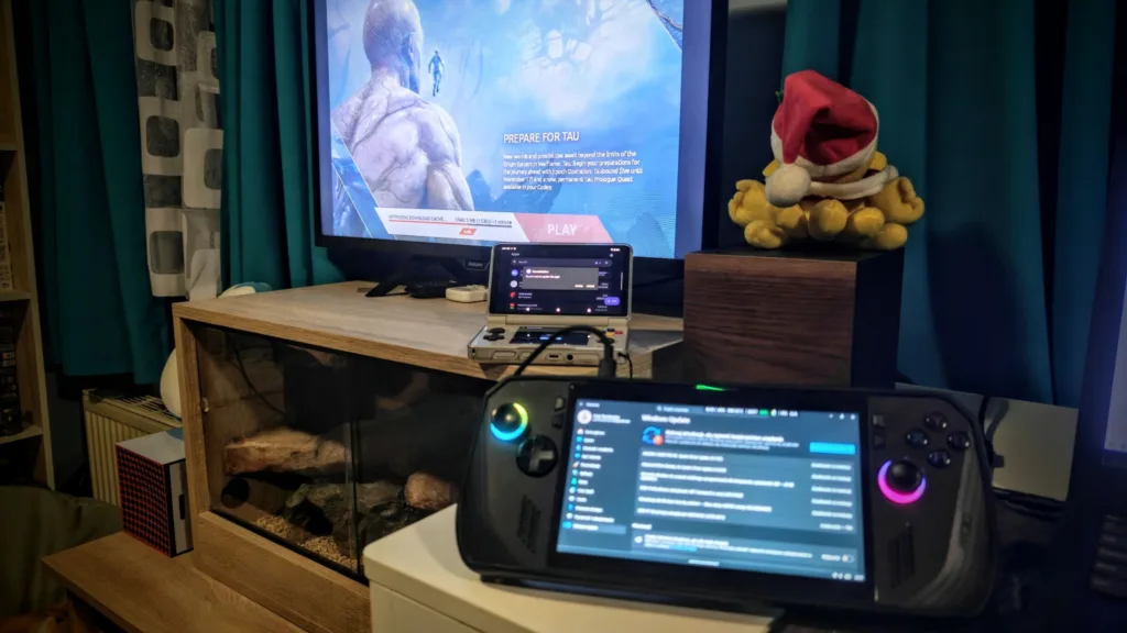 Ekran telewizora z grą Warframe, przenośna konsola do gier i mała pluszowa maskotka w czapce Mikołaja na drewnianej szafce.