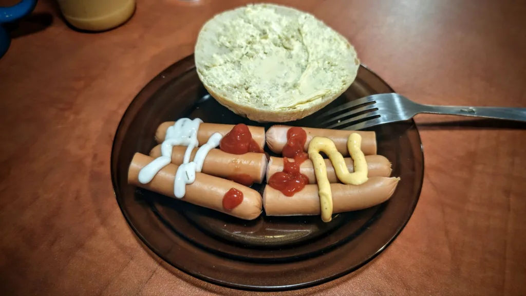 Na brązowym talerzu leżą cztery parówki polane majonezem, keczupem i musztardą, obok znajduje się przekrojona bułka posmarowana masłem oraz metalowy widelczykowy.