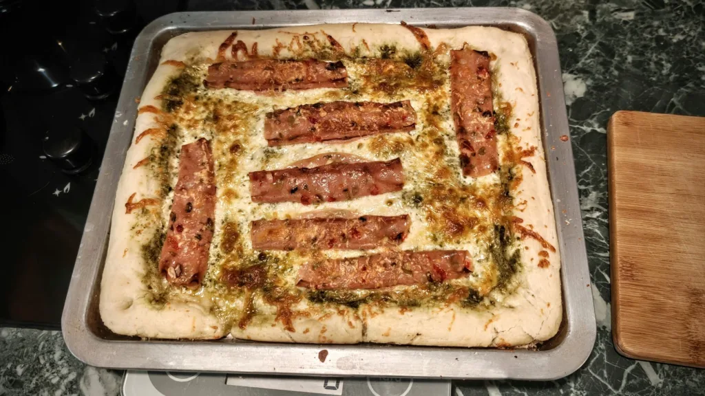 Domowa pizza na prostokątnej blasze z serem, zielonym sosem pesto i ułożonymi w rzędach kawałkami wędliny.