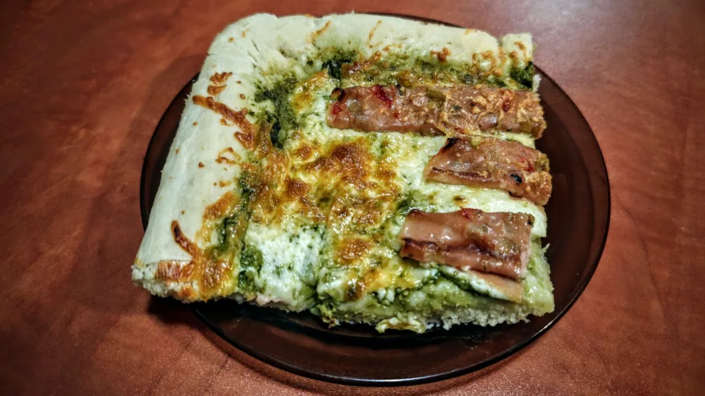 Kawałek domowej pizzy na ciemnym talerzu, pokryty zielonym pesto, topionym serem i trzema podłużnymi kawałkami wędliny.